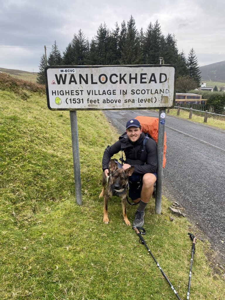 Nick und Nero gebeugt und in die Kamera lachend vor dem Ortsschild von Wanlockhead während der JOGLE Wanderung