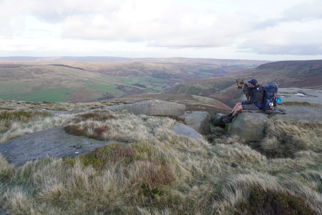 Nick und Nero sitzend auf einem Steinvorsprung mit einer Aussicht auf die hügelige Landschaft des Pennine Way während der JOGLE Wanderung